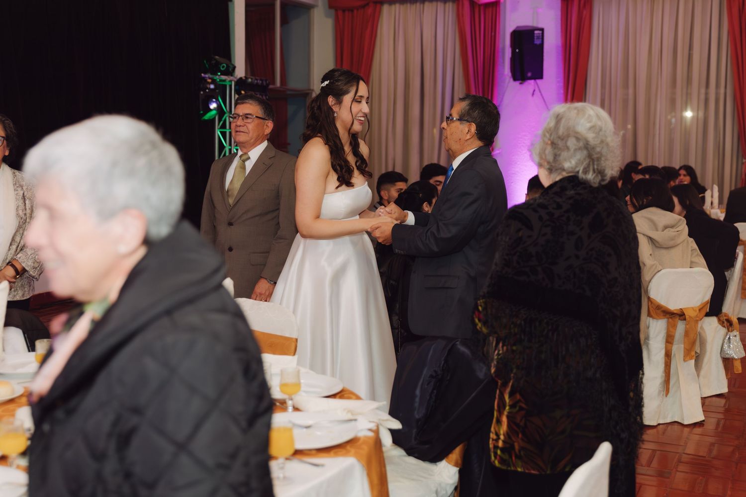 Bride in white gown greets elderly guests at elegant wedding reception hall.