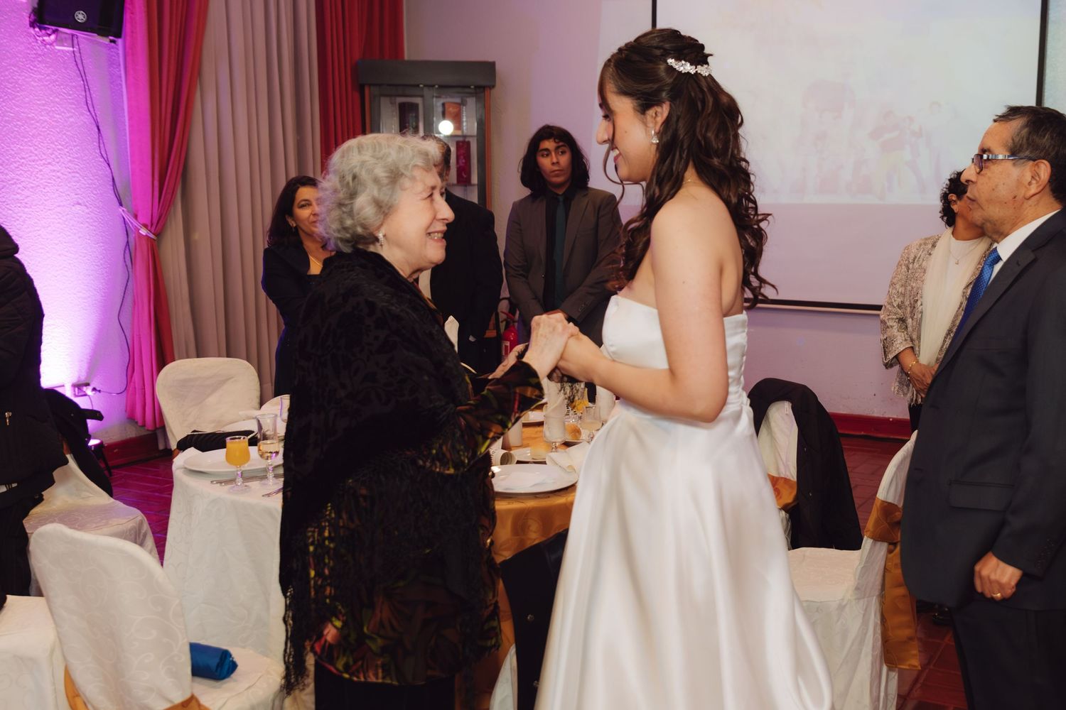 Bride in white wedding dress holds hands with elderly woman at reception.
