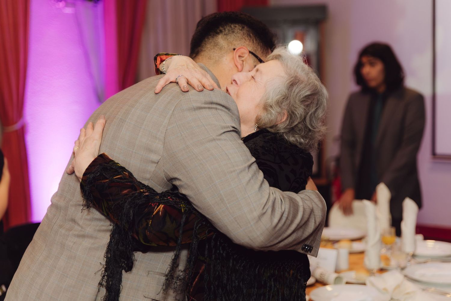 Man in grey suit shares tender hug with elderly woman at wedding.