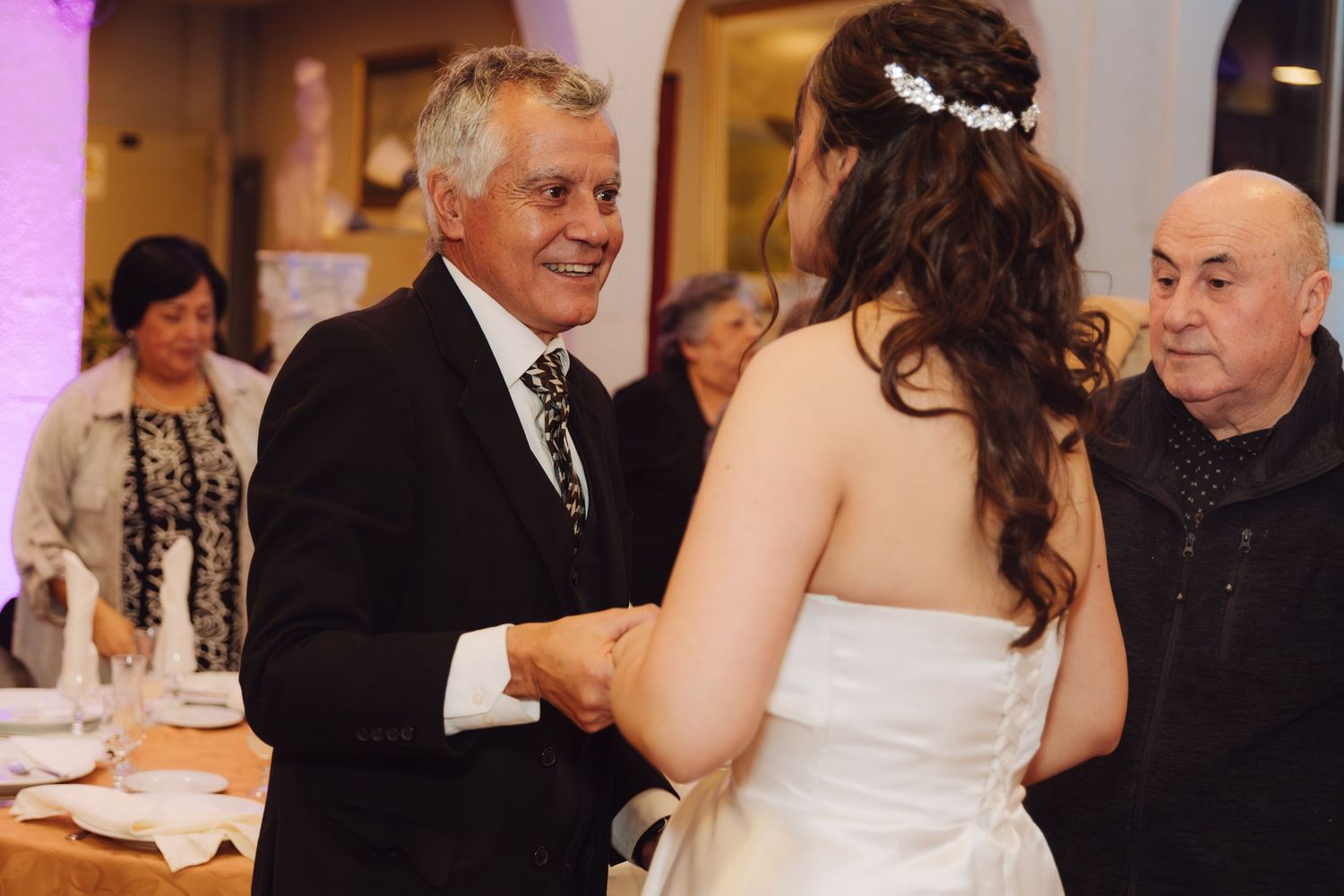 Smiling older man in suit shaking hands with bride in white gown at wedding reception.