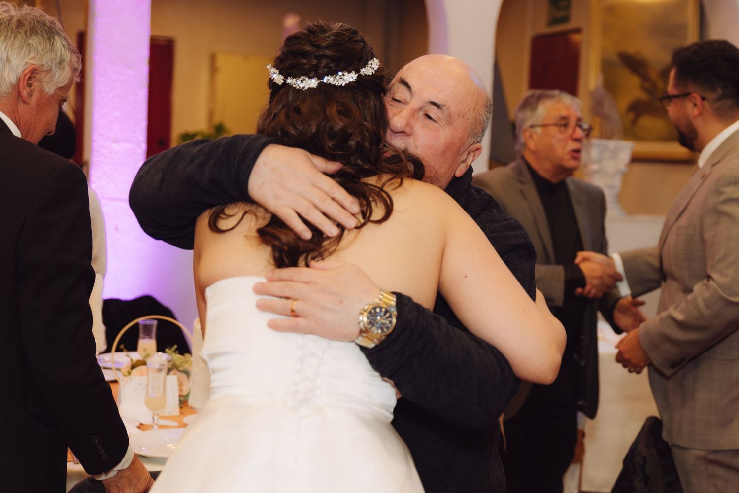 Bald man wearing gold watch embracing bride in white wedding dress at reception.