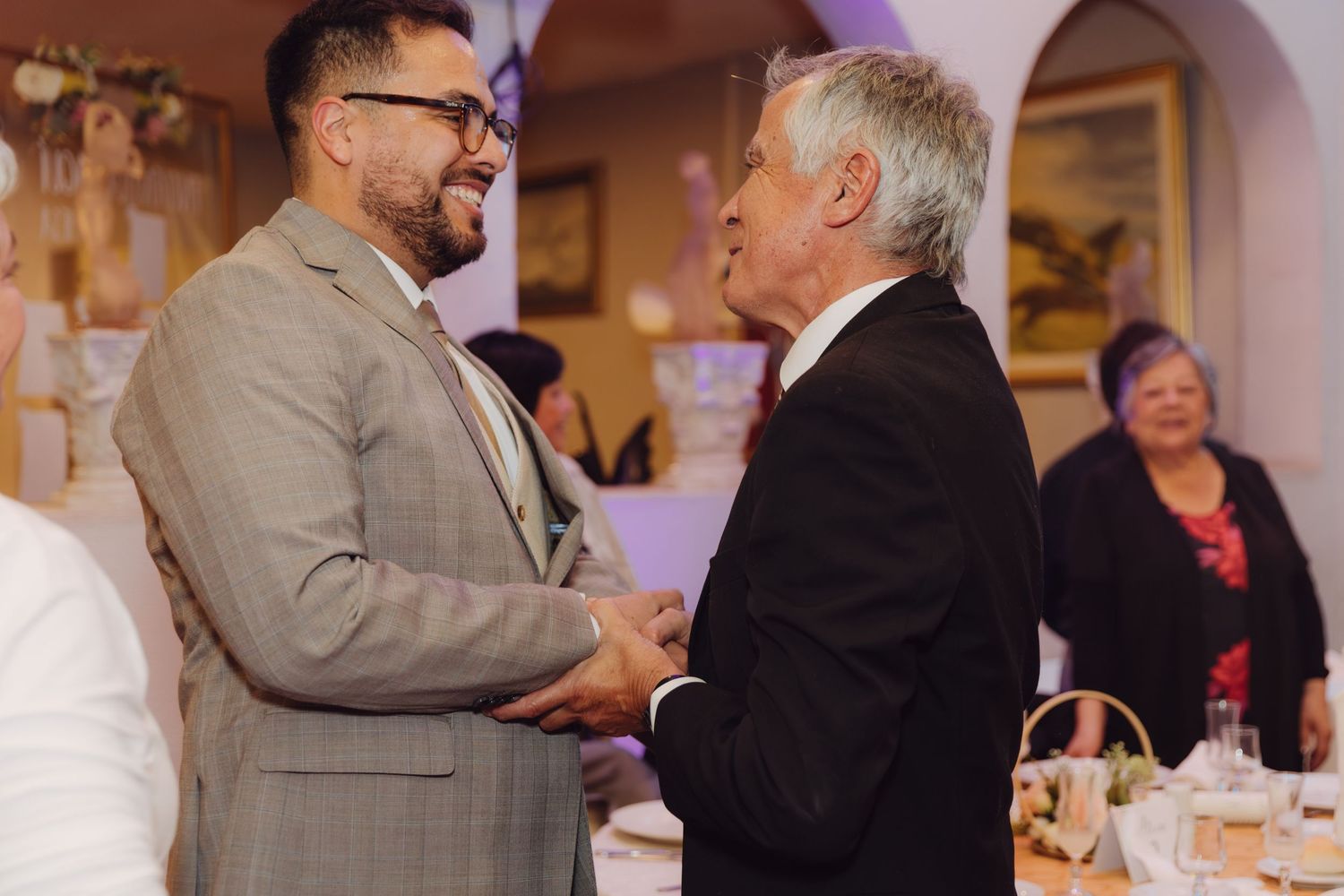 Two men in suits sharing a warm handshake and smiling at a wedding reception.