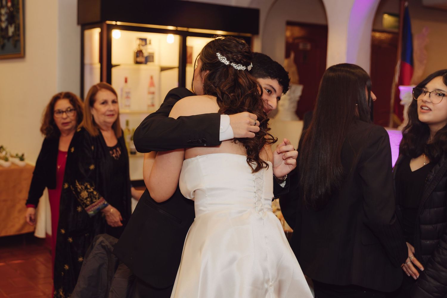 Bride in white gown receiving warm hugs from guests at her wedding reception.