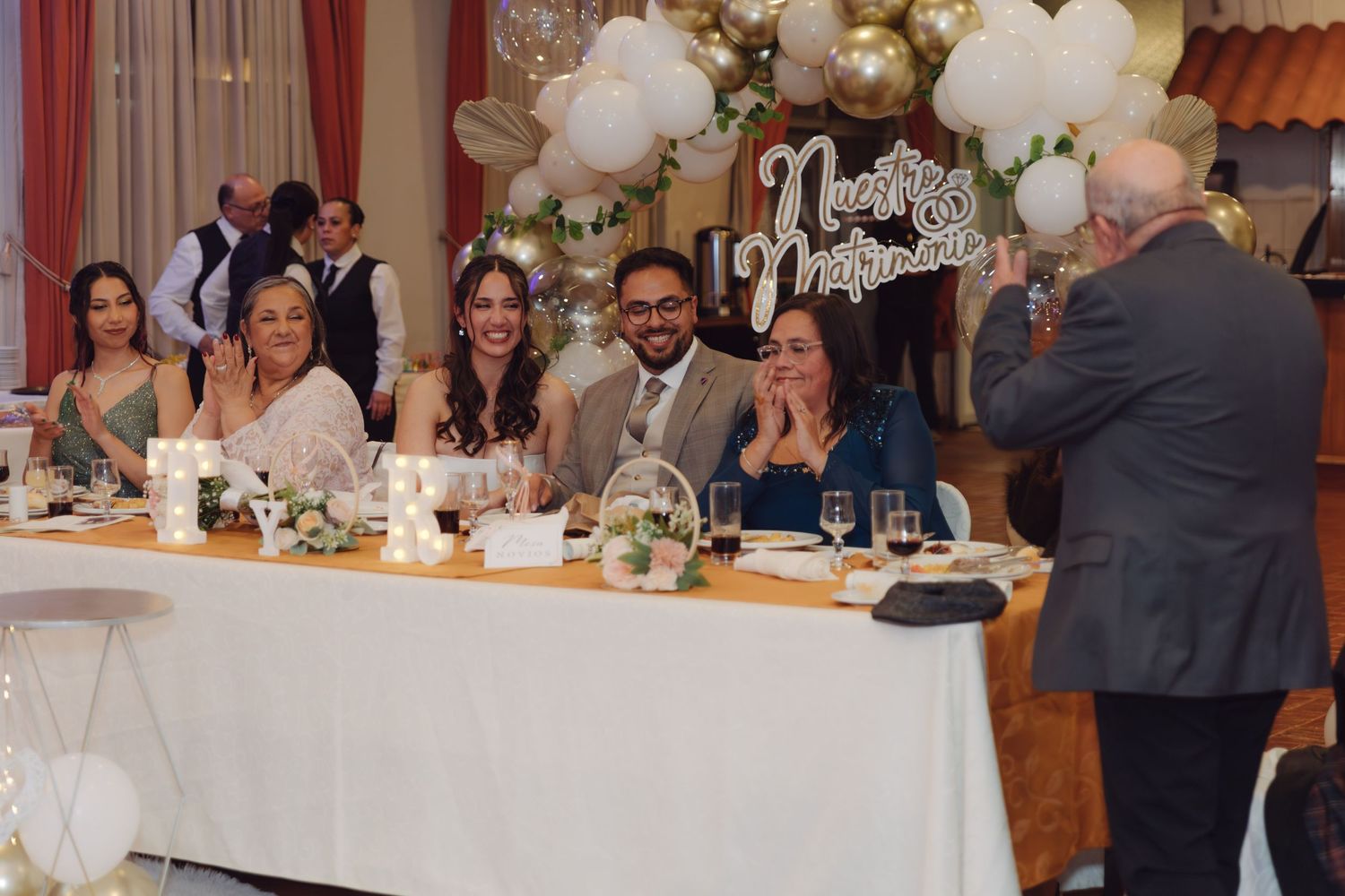 Wedding party laughing at head table decorated with white and gold balloons during reception.