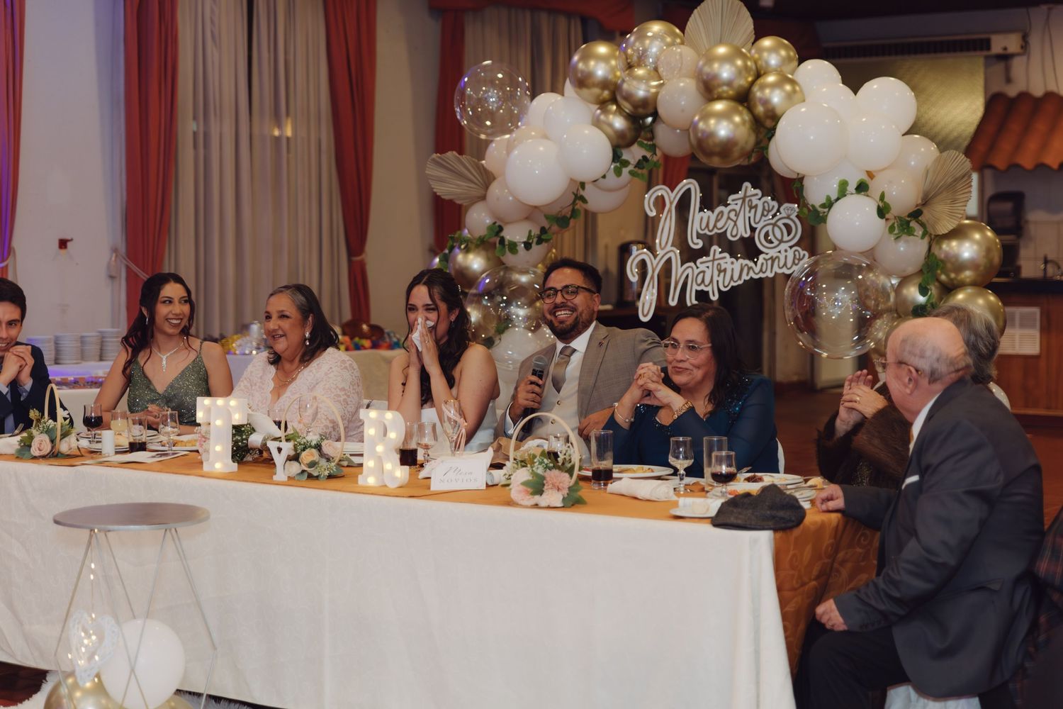 Bride and groom smile with family at elegant wedding reception head table with balloon arch.