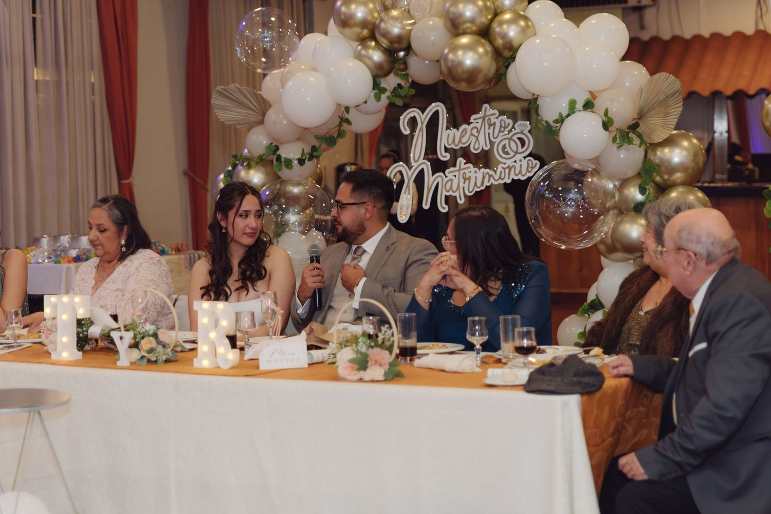 Groom speaks at wedding head table surrounded by family beneath white and gold balloon arch.