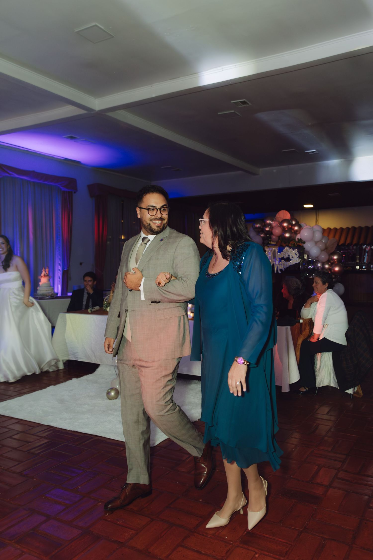 Groom in tan suit dances with woman in teal dress on wedding reception dance floor.
