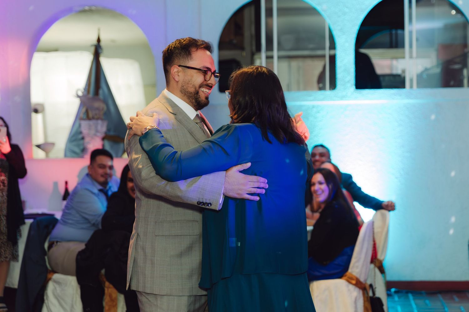 Groom in gray suit shares warm embrace with woman in blue dress at wedding reception.