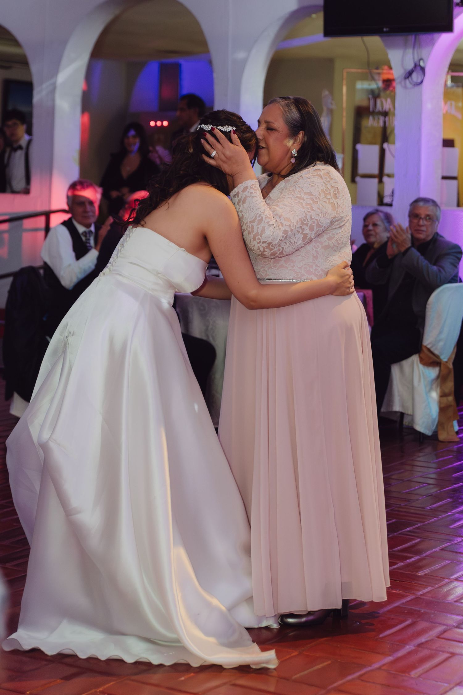 Bride in white gown shares emotional dance with mother in pink dress at wedding reception.