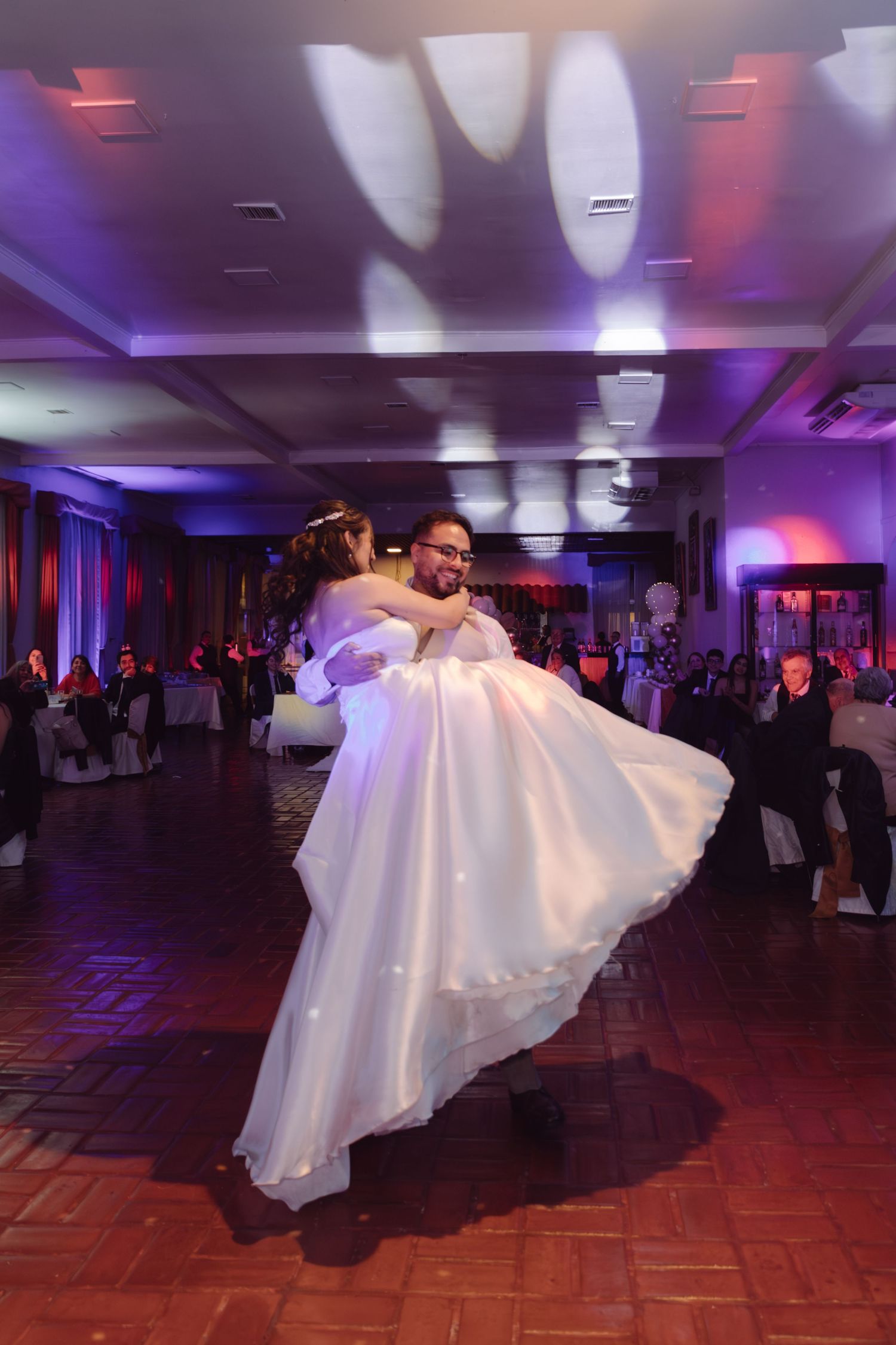 Groom lifts bride in white gown during romantic first dance at elegant wedding reception with purple lights.