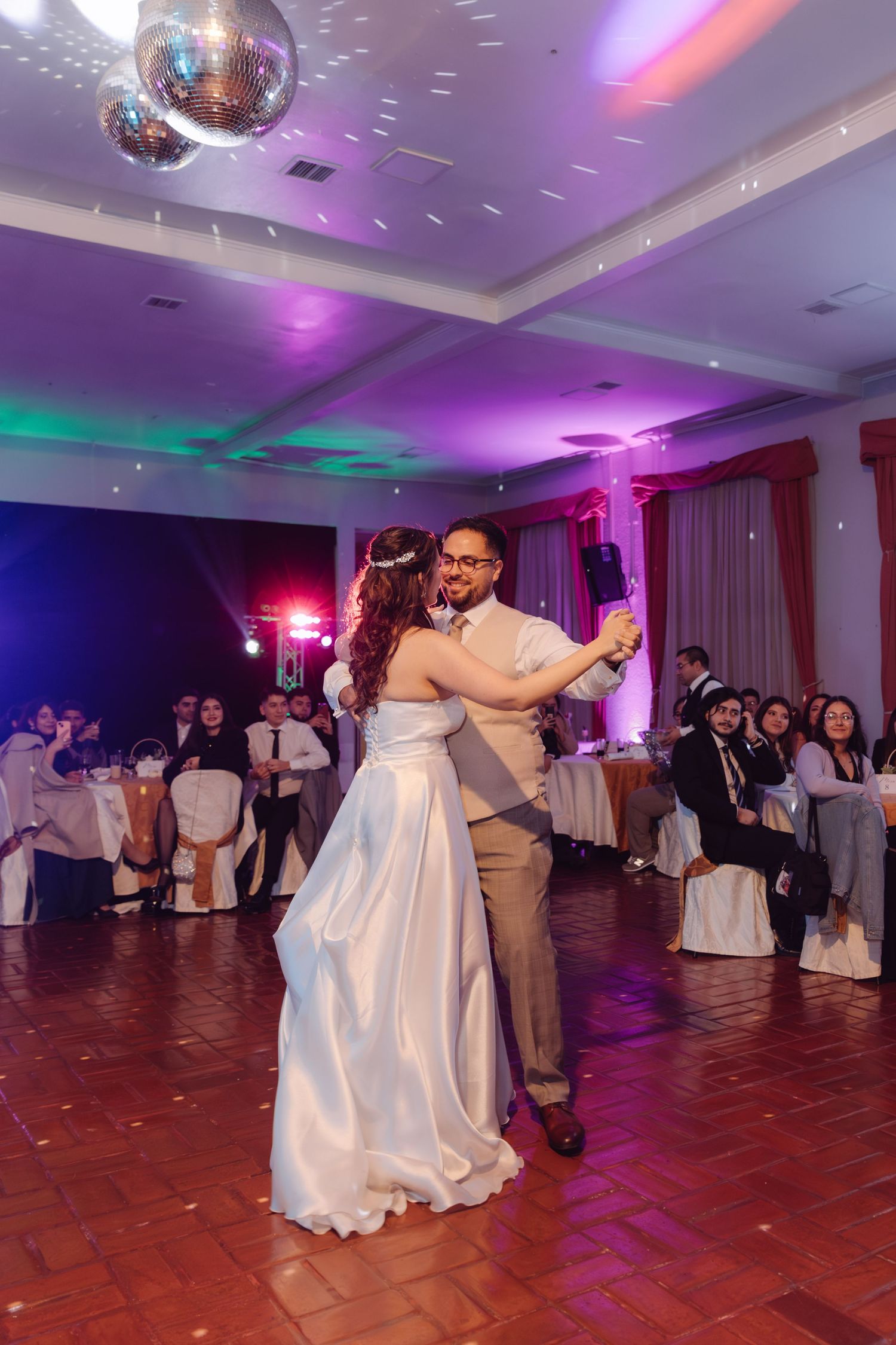 Bride and groom share first dance under disco ball at wedding reception with colorful ambient lighting.