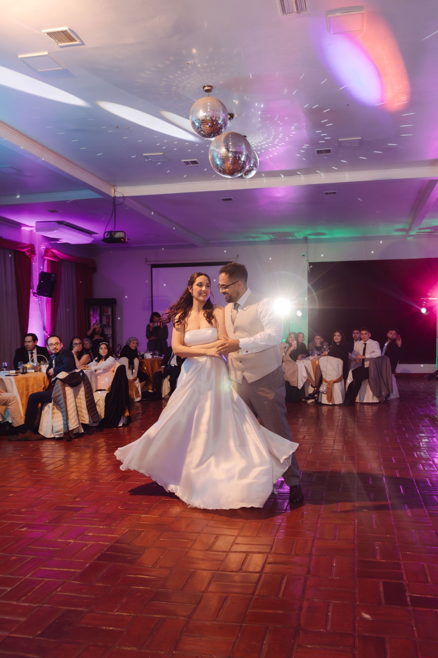 Newlyweds dance elegantly under disco balls at wedding reception with purple and green ambient lighting.