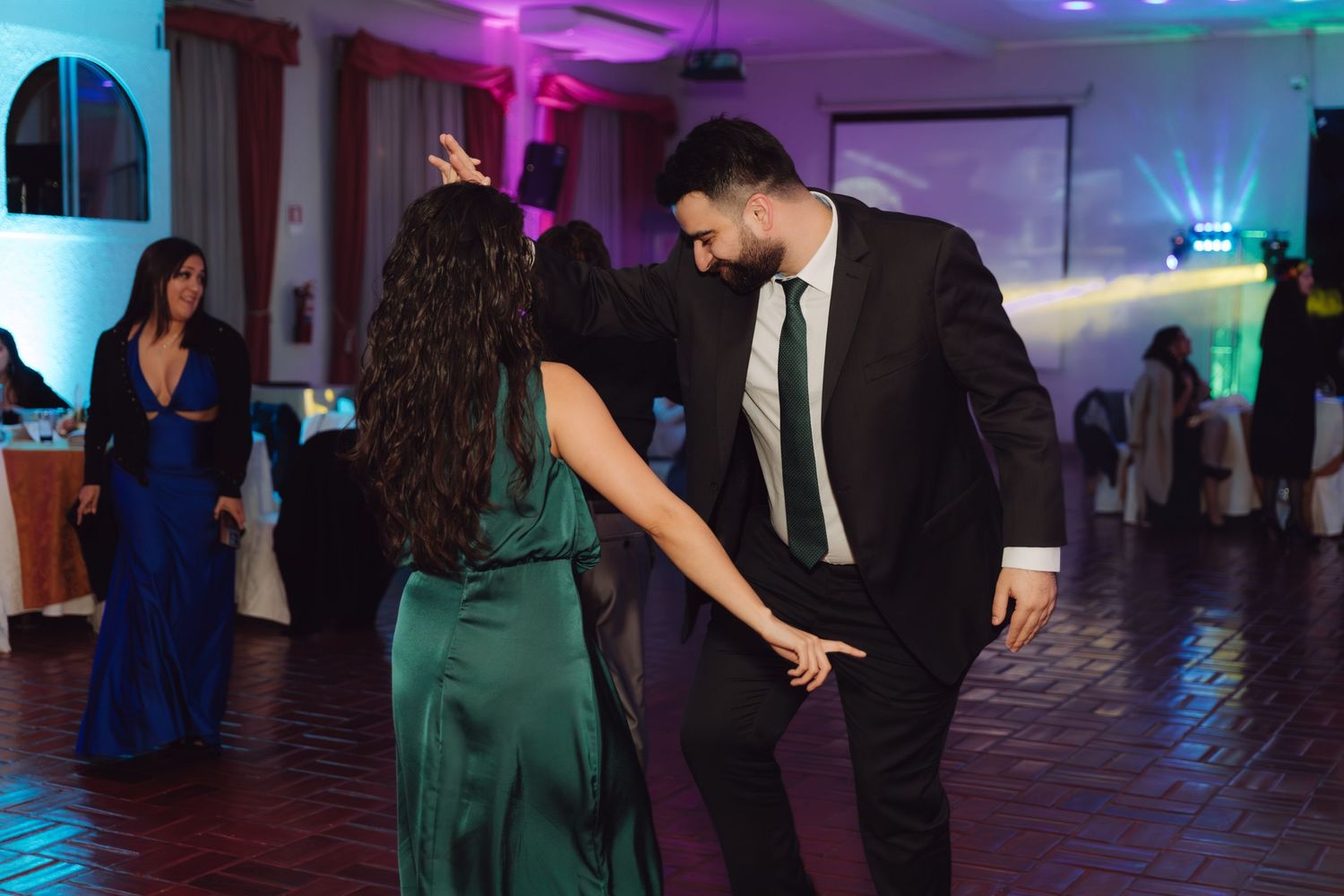 Man in black suit dances with woman in green satin dress at lively wedding reception.