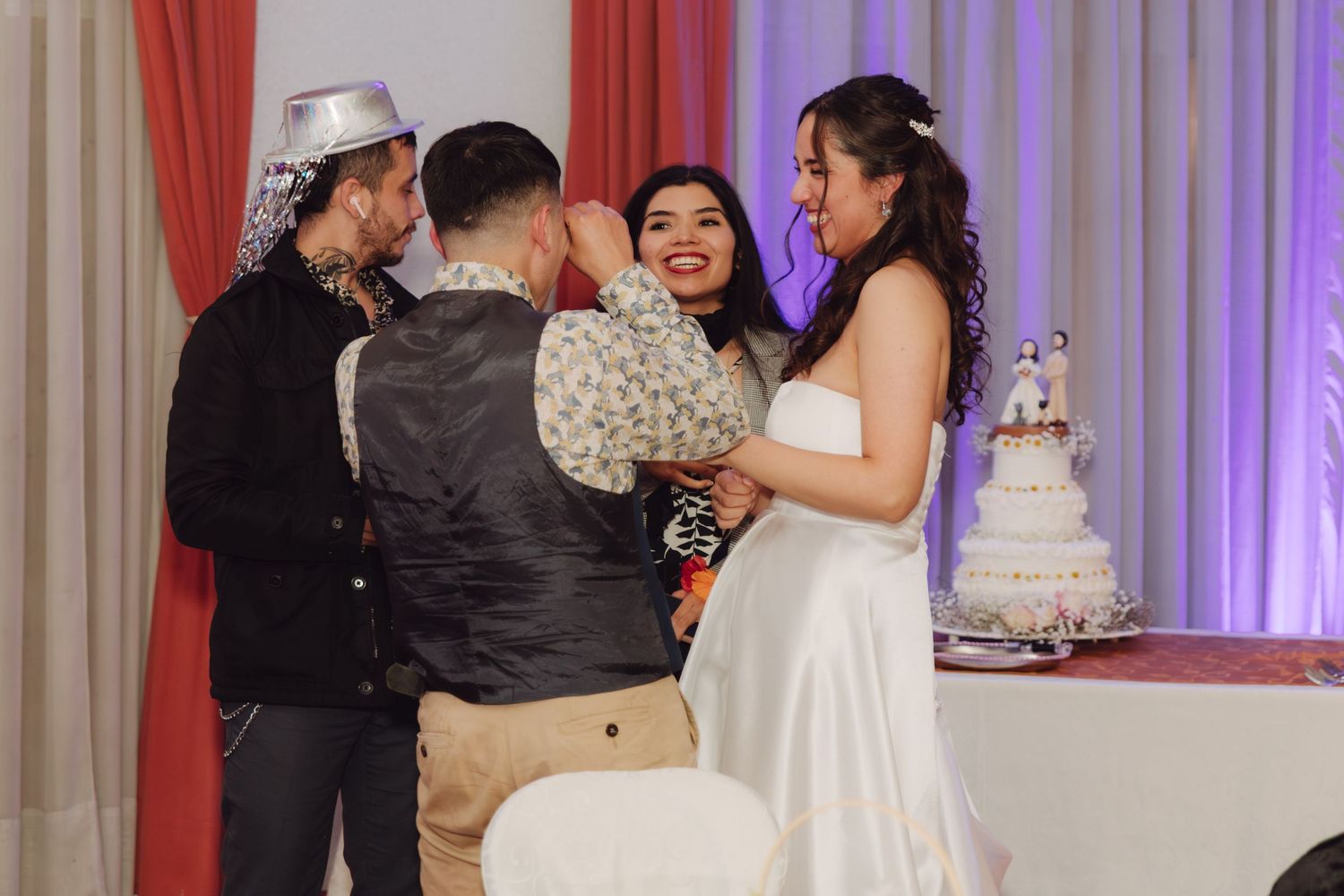 Bride in white gown smiles with guests near tiered wedding cake at reception celebration.
