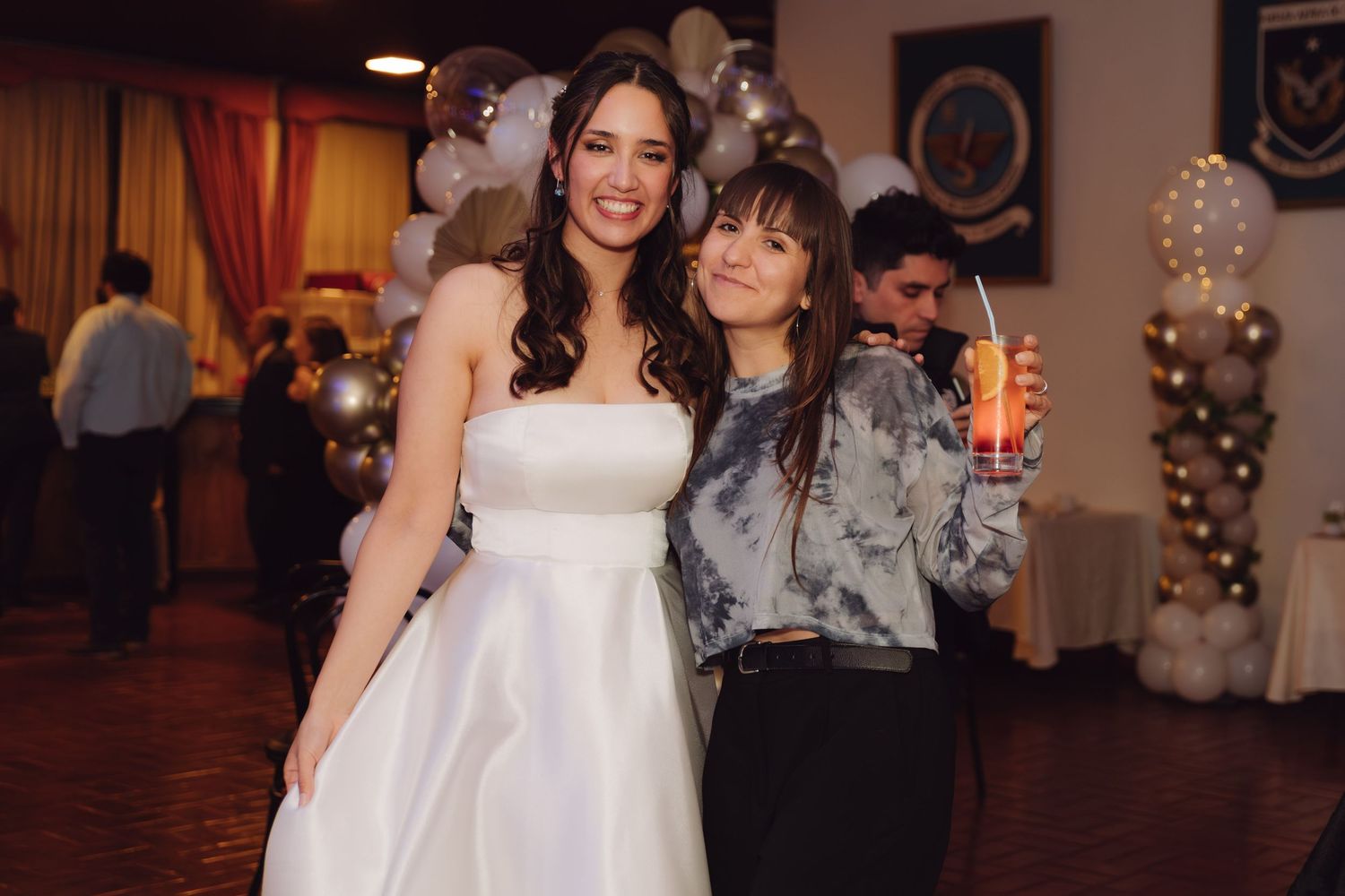 Bride in white gown poses with smiling female guest holding a drink at wedding reception.