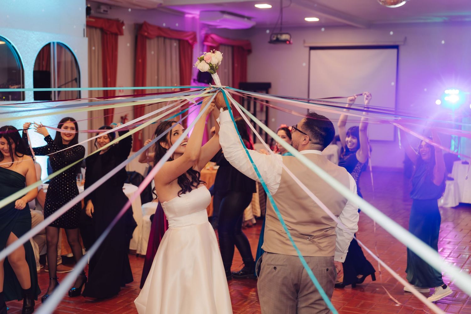 Bride and groom hold bouquet with colorful ribbons stretched to wedding guests on dance floor.
