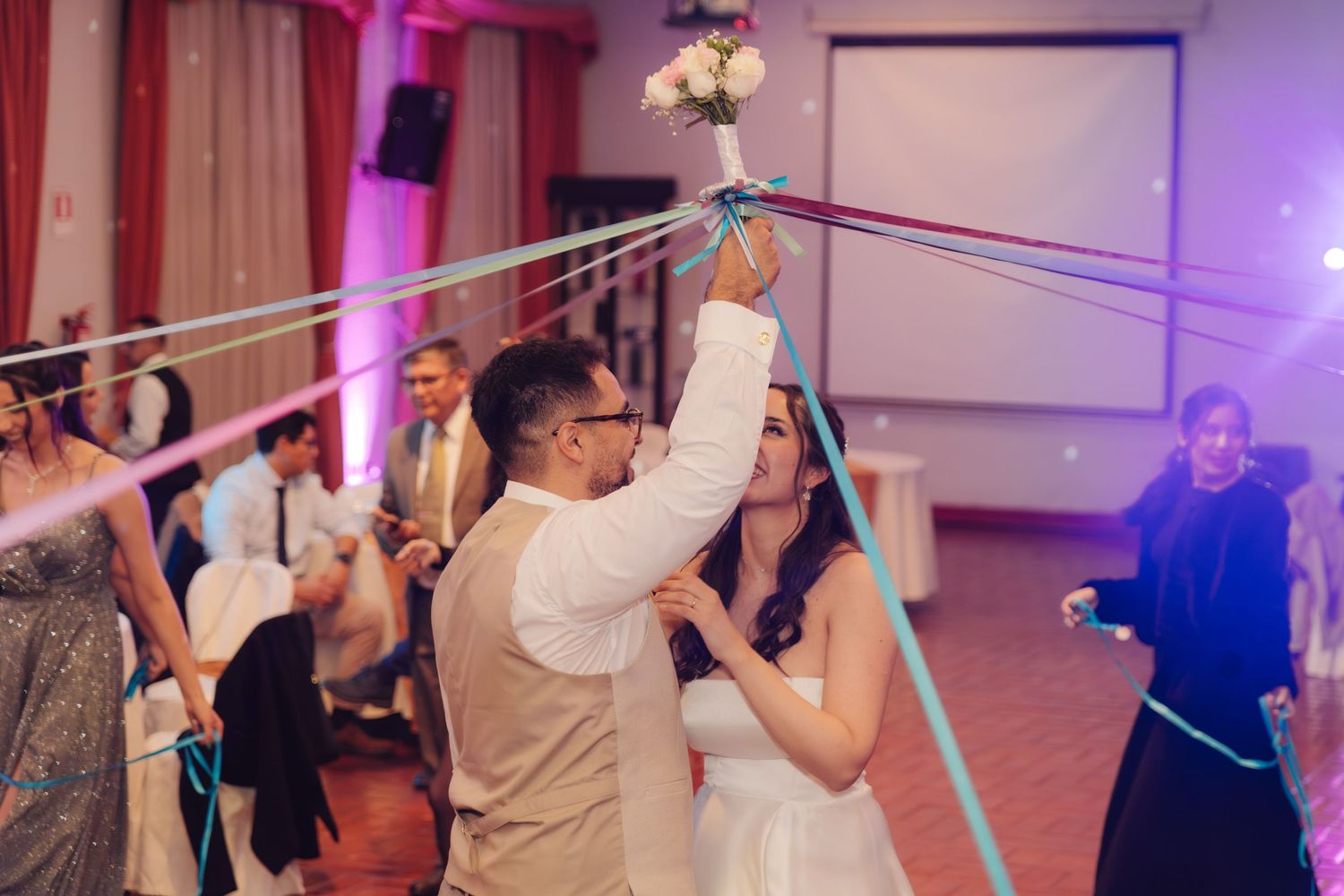 Groom in vest holds bridal bouquet with colorful streamers during wedding reception dance.