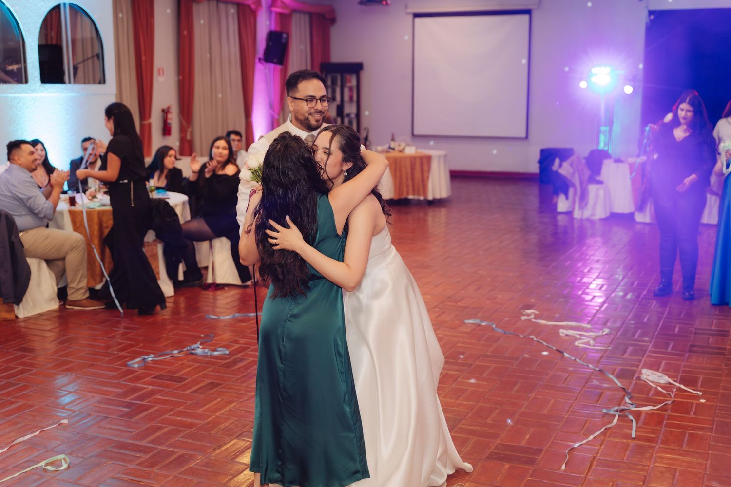 Bride in white gown embraces woman in green dress during emotional wedding reception dance.