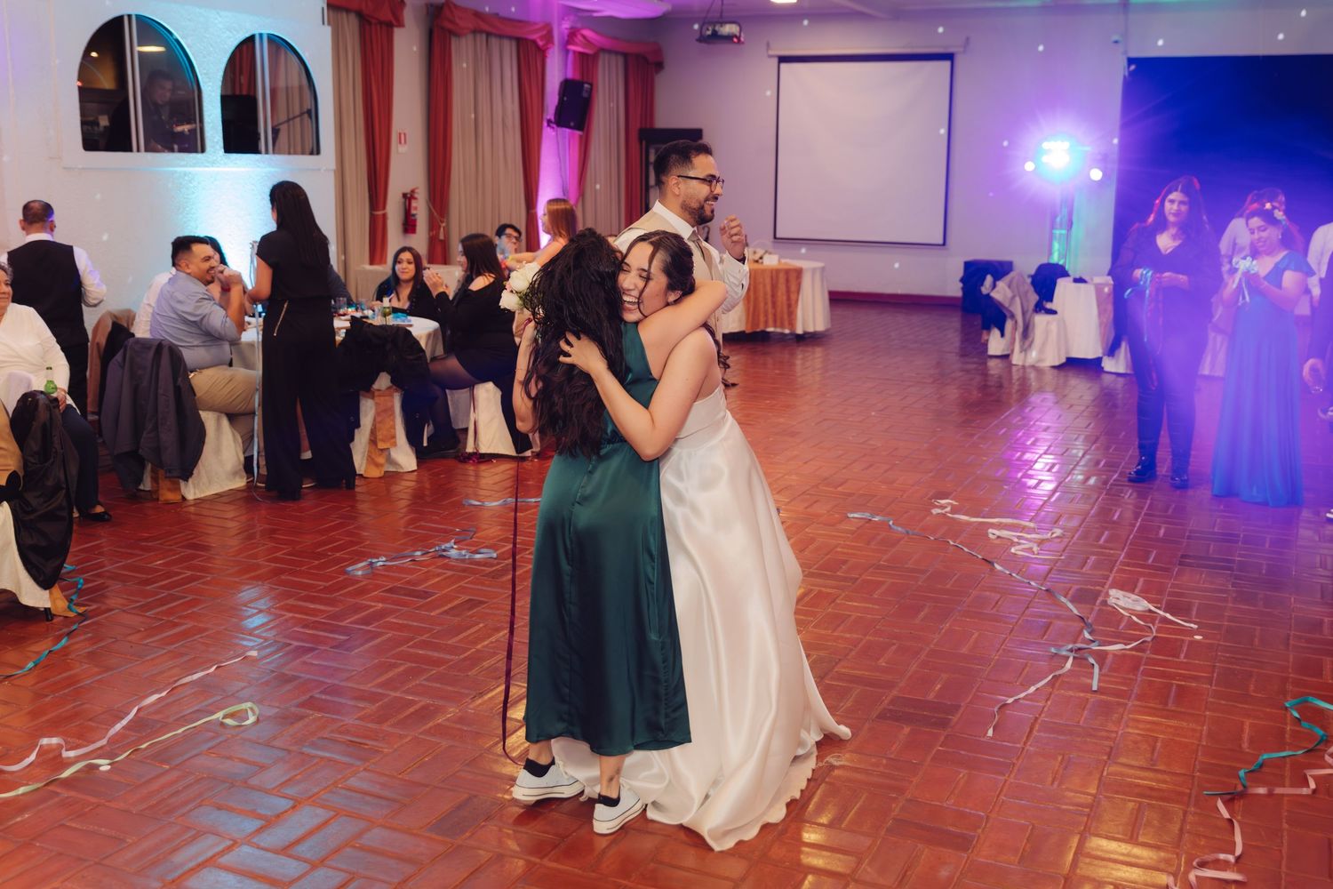 Bride hugs woman in emerald green dress on dance floor at elegant wedding reception.