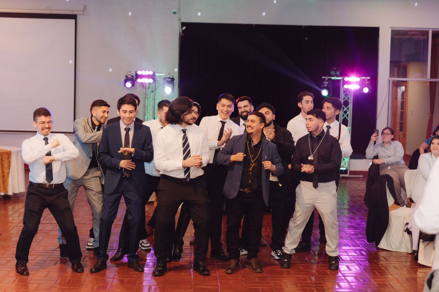 Group of men in suits pose playfully on dance floor at wedding reception celebration.