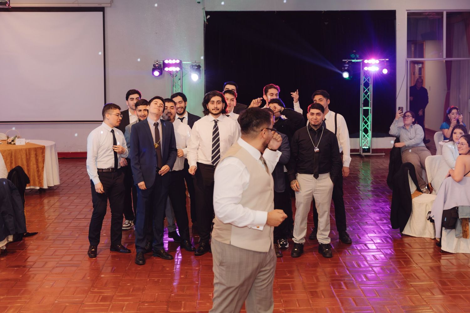 Groom in white vest prepares to toss garter to group of men at wedding.