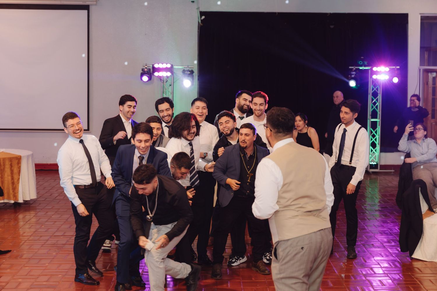 Group of groomsmen in suits and suspenders celebrating at a wedding reception on the dance floor.