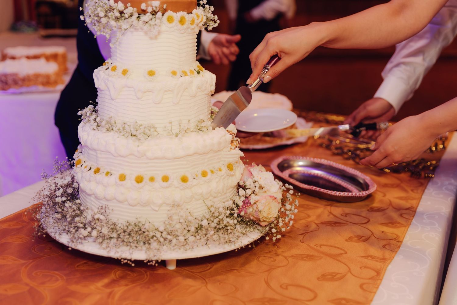 Close-up of elegant three-tier white wedding cake with yellow accents being cut at reception.