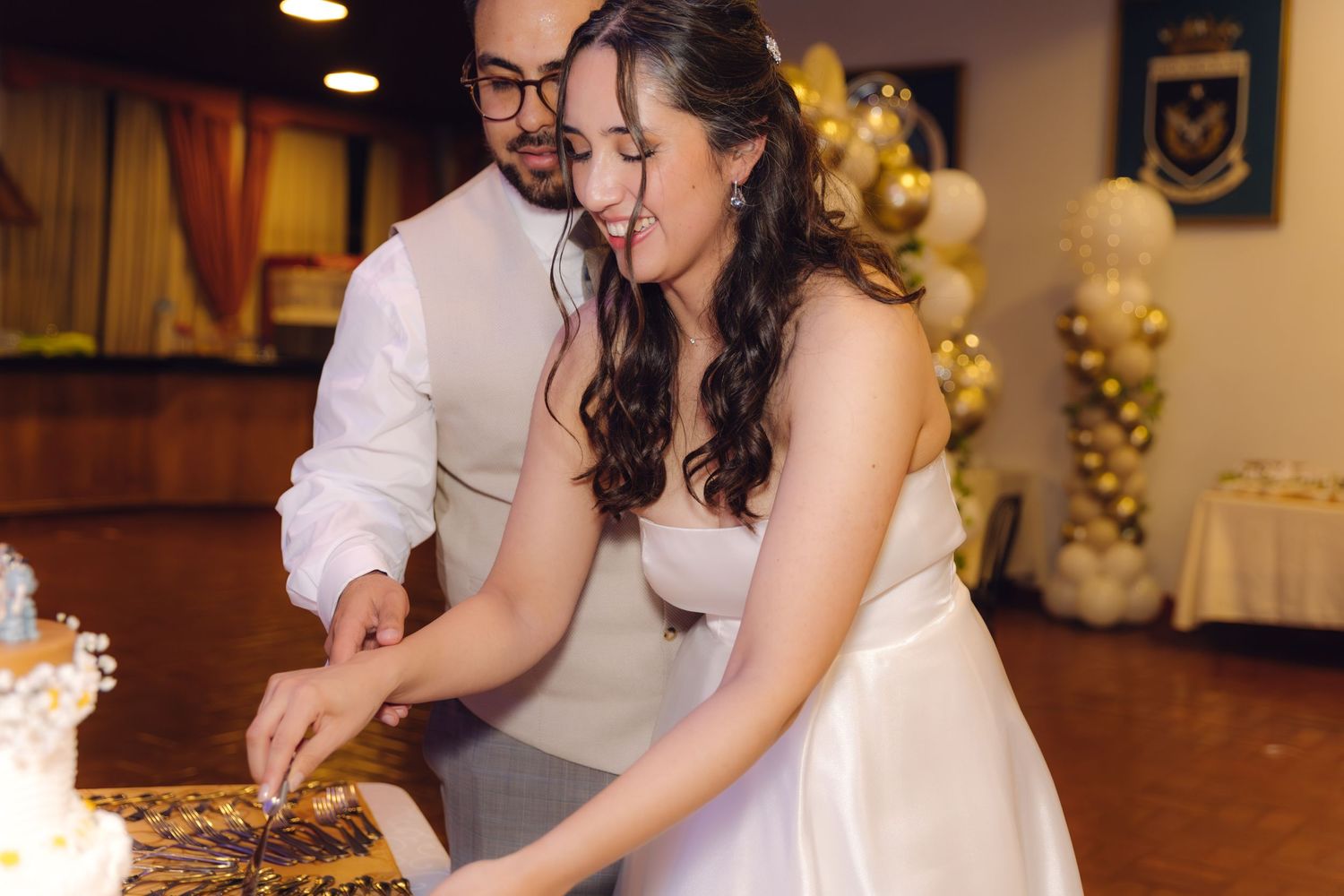 Bride and groom smiling while cutting their decorated wedding cake at reception venue.