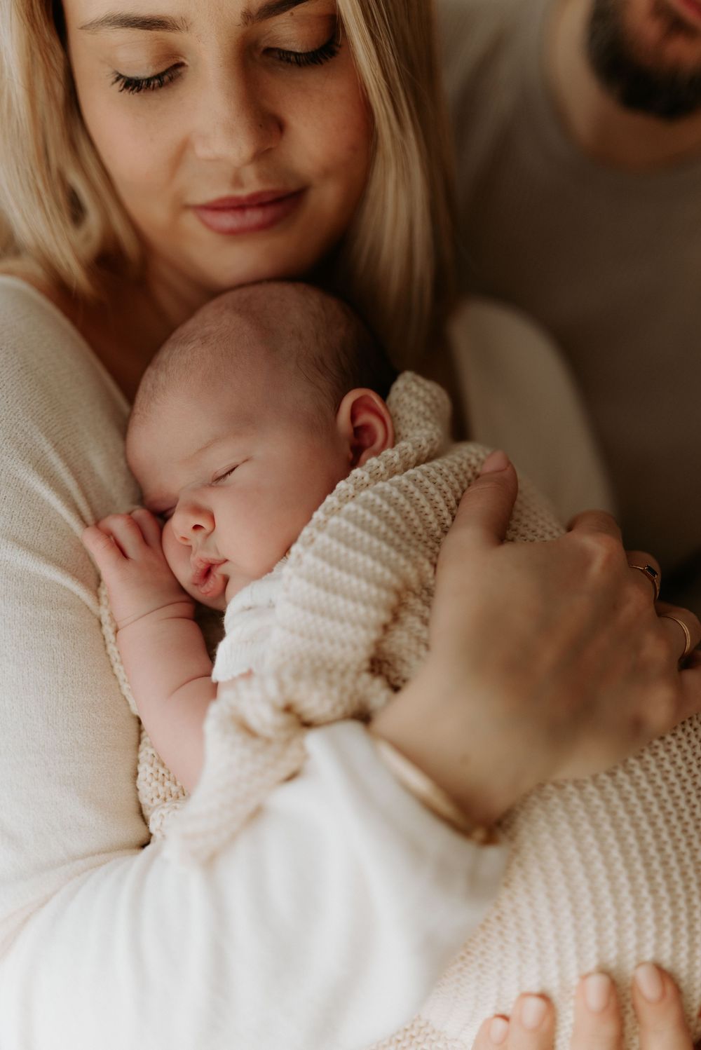 A sleeping newborn baby wrapped in a cozy white knit sweater rests peacefully against someone in soft lighting.