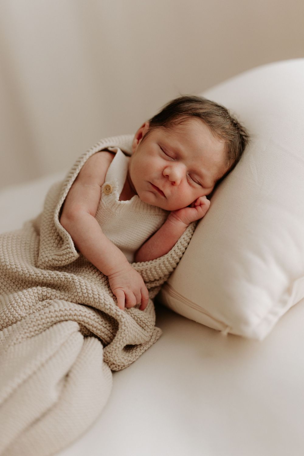 A sleeping newborn baby wrapped in a cozy beige knit blanket lies peacefully on white bedding.