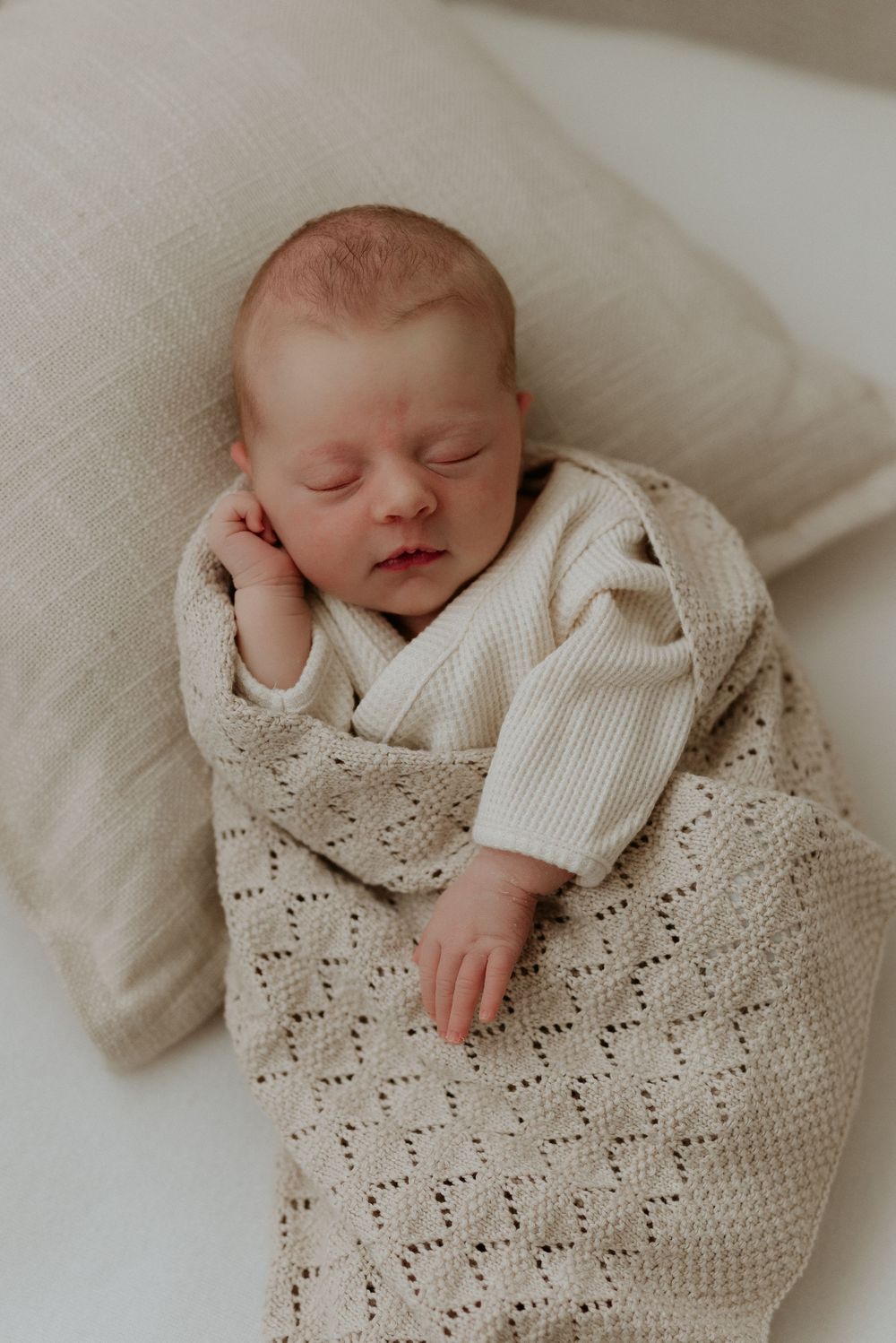 Peaceful newborn baby sleeping wrapped in a soft cream knit blanket on a white pillow.