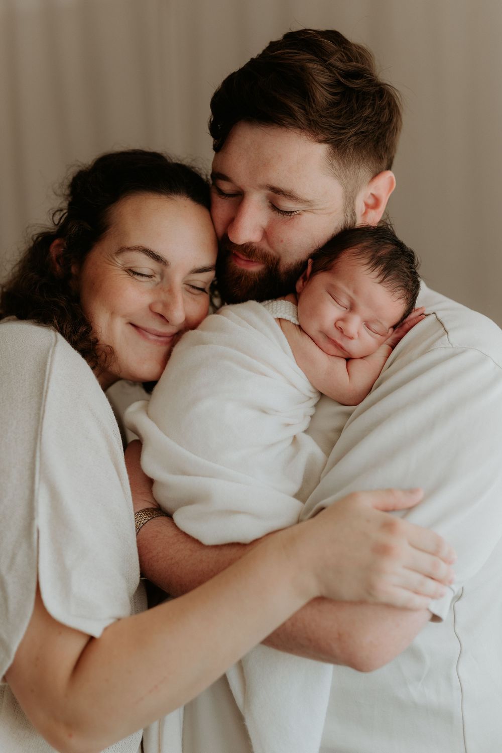 New parents tenderly embrace their sleeping newborn baby wrapped in white, sharing a peaceful family moment.