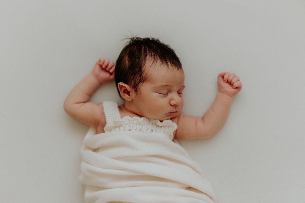 Peaceful newborn baby sleeping on white background, wrapped in cream swaddle with arms raised, dark hair visible.