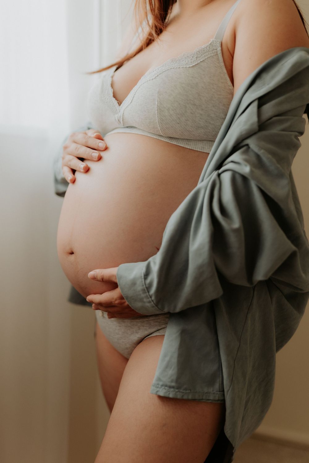 Pregnant woman in gray lingerie and open shirt cradling her baby bump near a bright window.