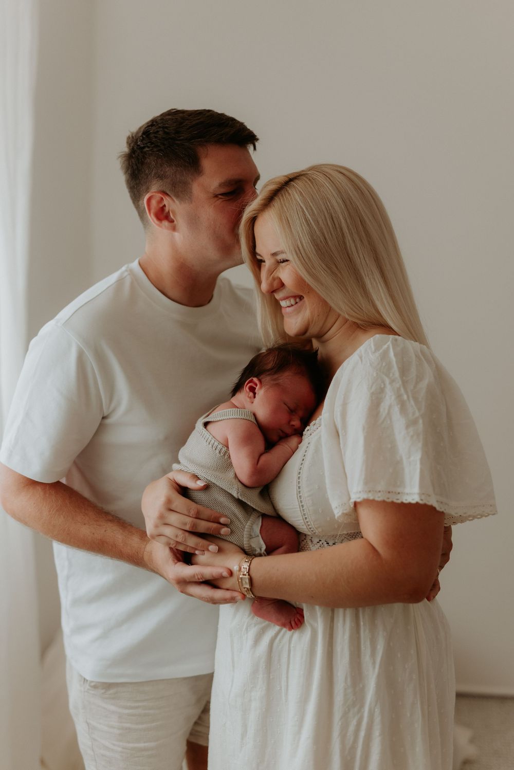 A couple in white outfits smiles while tenderly holding their newborn baby in a bright, airy room.