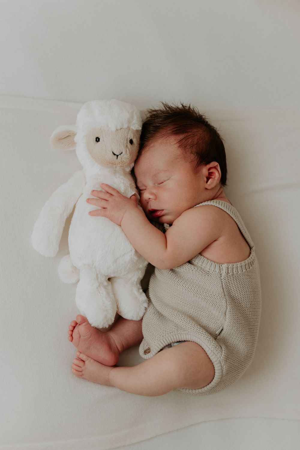 Newborn baby sleeping peacefully in a beige knit romper while cuddling a white lamb stuffed animal on a white blanket.