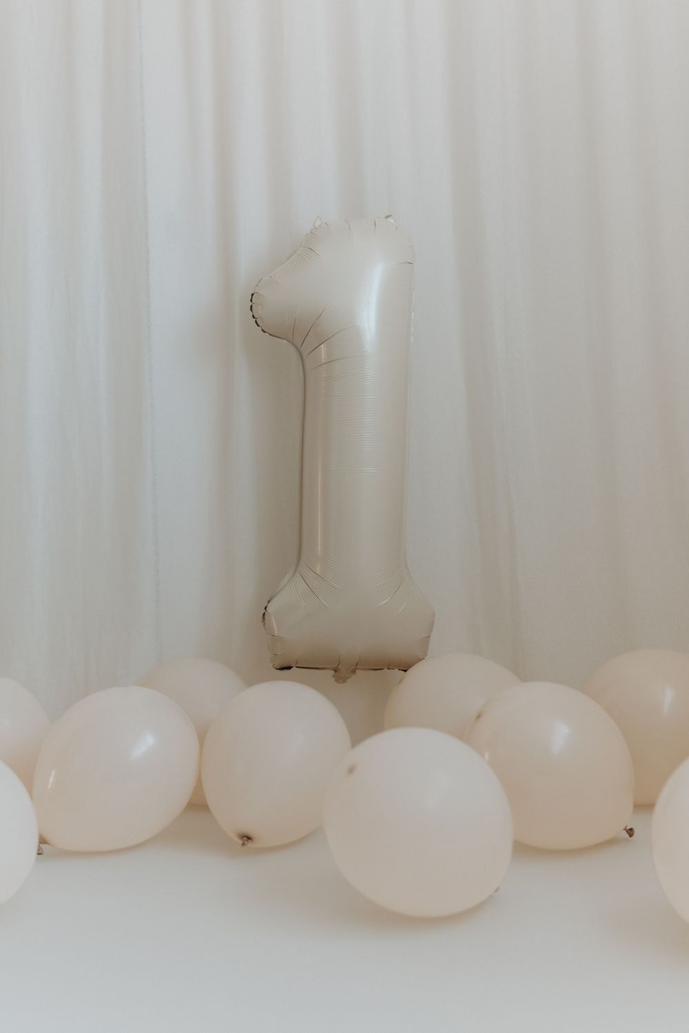 A large number one foil balloon surrounded by small white latex balloons against a white curtain backdrop.