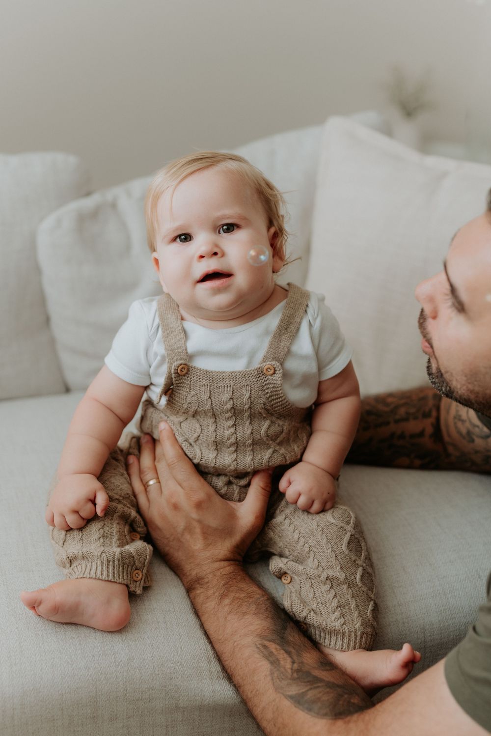 A tattooed father holds his smiling baby dressed in a knit tan overalls on a white couch.