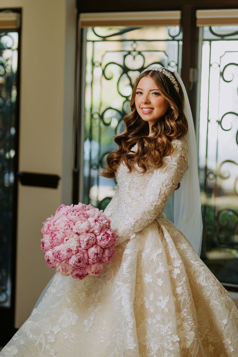 A stunning bride in a white lace gown with long sleeves poses with a pink peony bouquet against an elegant backdrop.