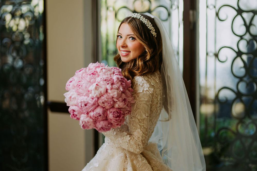 A bride in a lace wedding gown holds a large pink peony bouquet while standing by an ornate window.