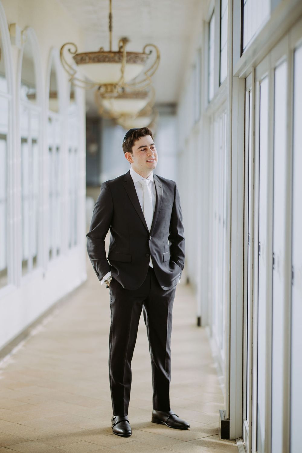 A groom in a classic black tuxedo poses in an elegant hallway with chandeliers.