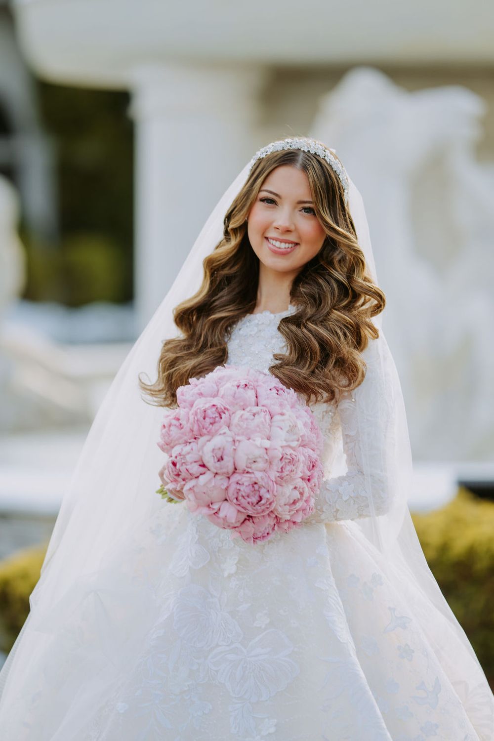 A bride in a white wedding gown holds a pink bouquet of roses while wearing a delicate veil.