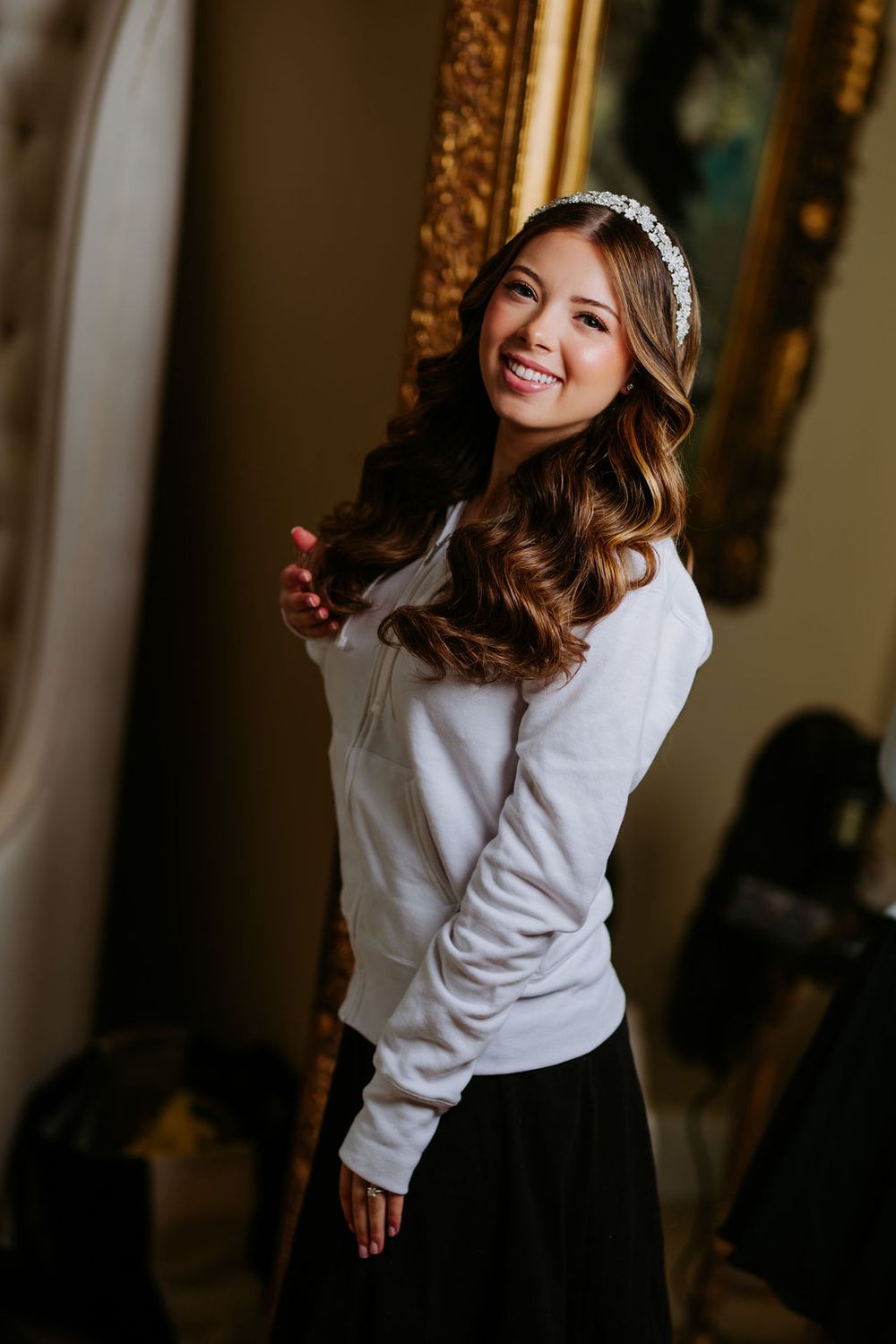 A joyful bride in a white sweater poses in an elegant indoor setting with ornate decor.