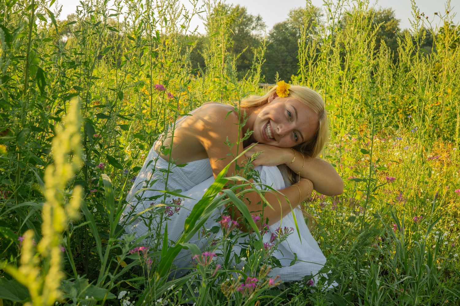 A girl in a flower field wears a long white dress and a flower in her hair as she smiles and laughs during her senior portraits in Michigan.