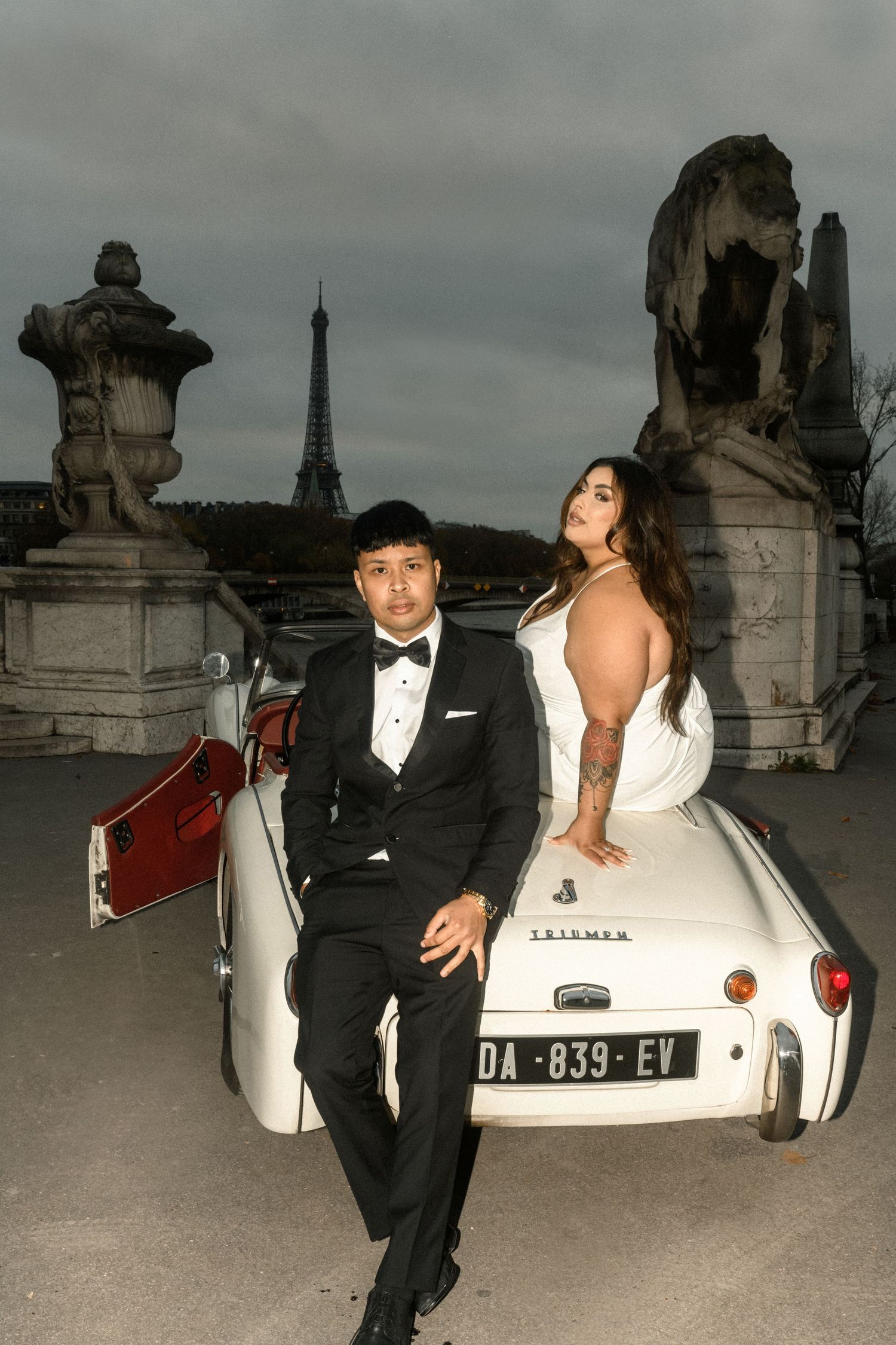 A glamorous wedding couple poses with a vintage car against a moody Parisian skyline with the Eiffel Tower visible.