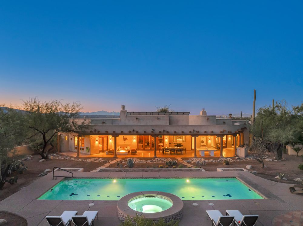 Panoramic view of a luxury desert home with illuminated pool and hot tub at dusk against a deep blue sky.