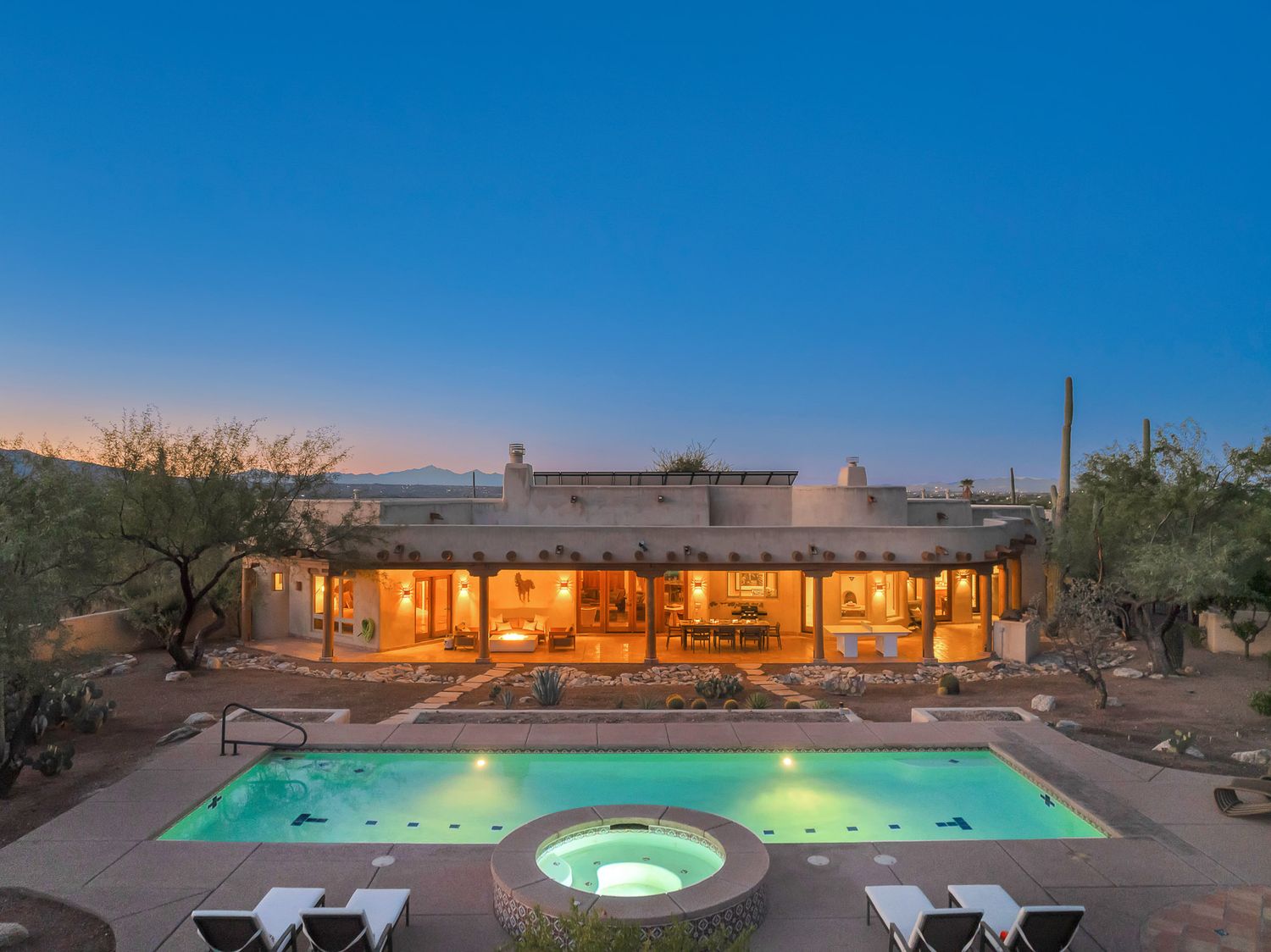 Panoramic view of a luxury desert home with illuminated pool and hot tub at dusk against a deep blue sky.