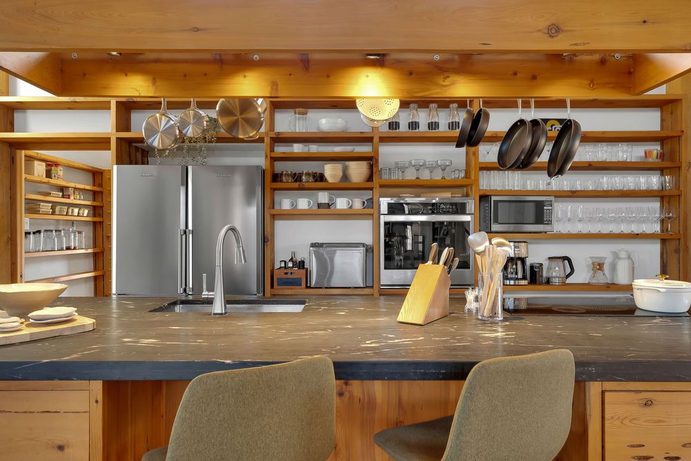 Modern kitchen interior with wood cabinets, stainless steel appliances, and open shelving displaying glassware.