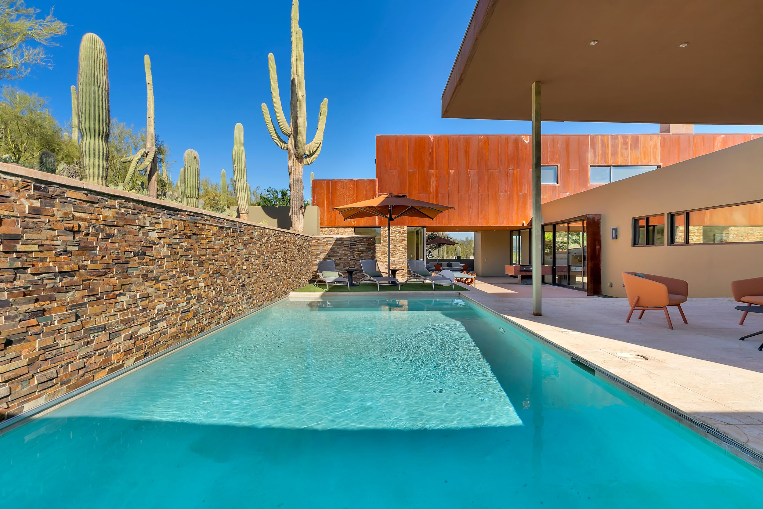 Stunning desert oasis pool surrounded by tall saguaro cacti and modern architecture with orange accents at sunset.