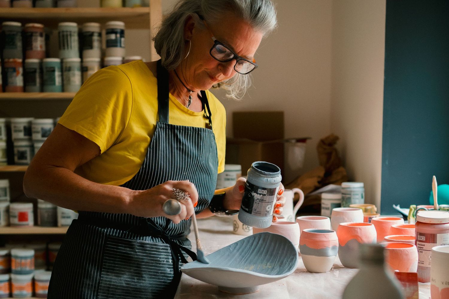 Artist in yellow shirt and green apron working with ceramic bowls in pottery studio with shelves of supplies.
